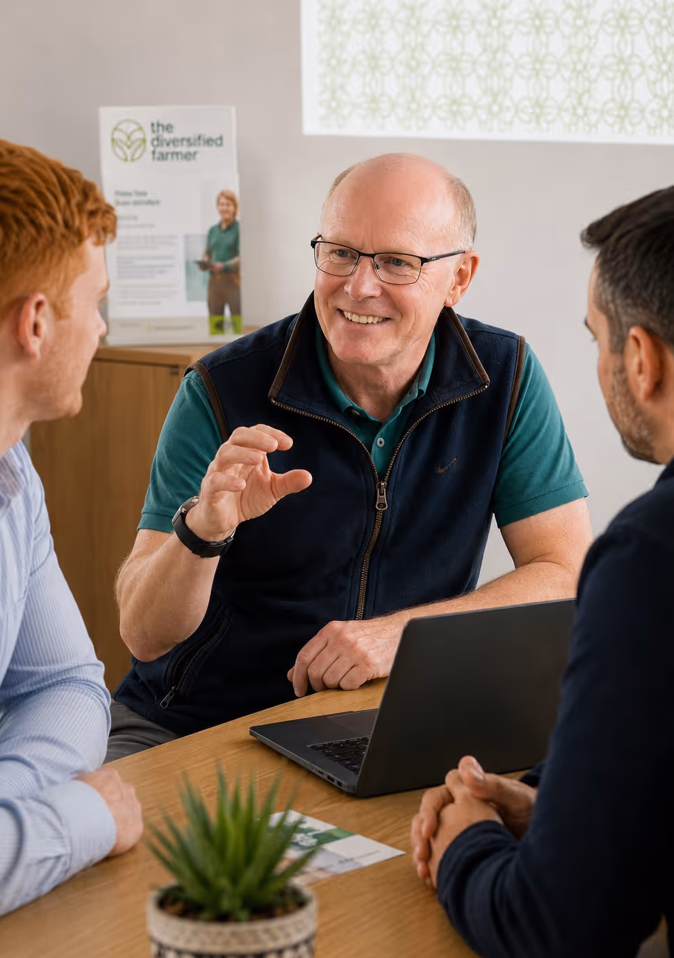 Emlyn explains farm diversification to two men seated at a table with a laptop and projection screen.